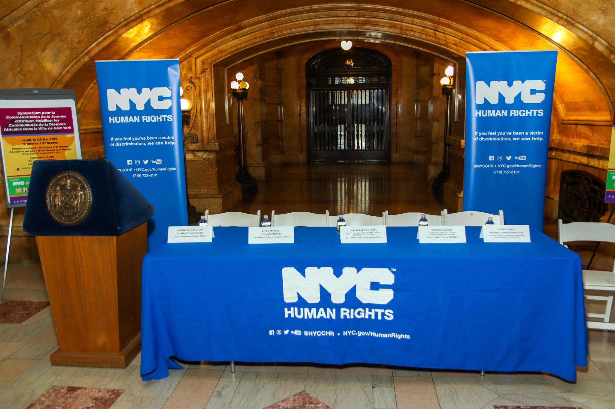 Photo of empty dais table with microphones, with background of event flyer (translated from French), “Africa Day Symposium: Engaging Communities of the African Diaspora in NYC."