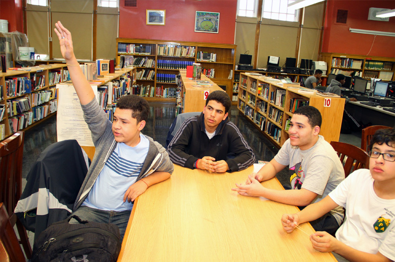 four male students at a table in the library with one boy raising his hand