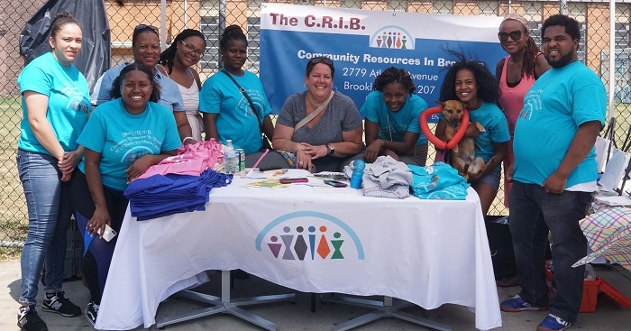 People surrounding a table of various items with The C.R.I.B Community Resources in Brooklyn sign in the background