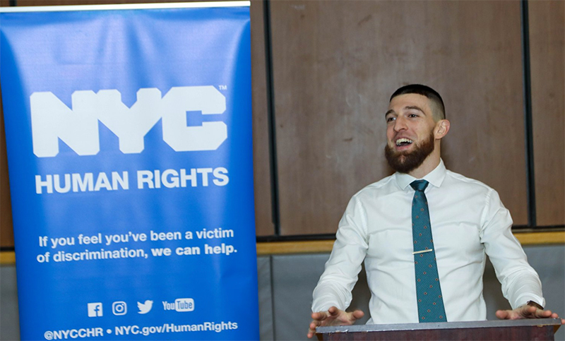 Commission employee speaking at a podium with blue banner behind him that reads in white lettering, "NYC Human Rights. If you feel you've been a victim of discrimination, we can help. @NYCCHR. NYC.gov/HumanRights."
