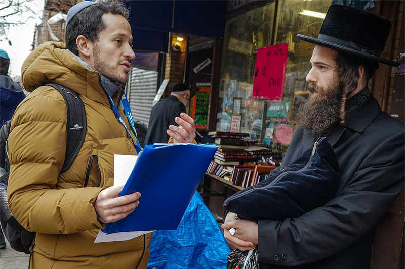 Man in yarmulke, with NYC Human Rights lanyard, on speaking with a man dressed in traditional Hasidic Jewish clothing, standing in front of a grocery store