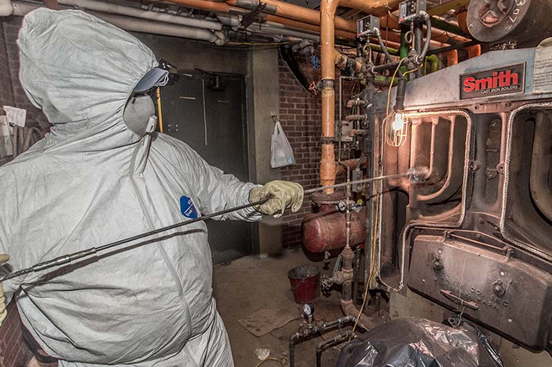 An employee wearing white protective gear, a face mask and gloves to perform maintenance on boiler room equipment.