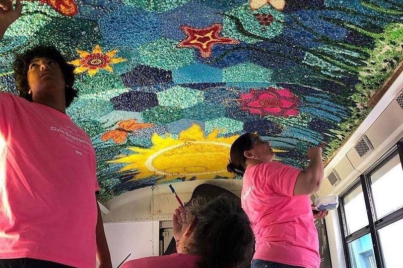 Three people with pink shirts install beads on the ceiling of The People's Bus.