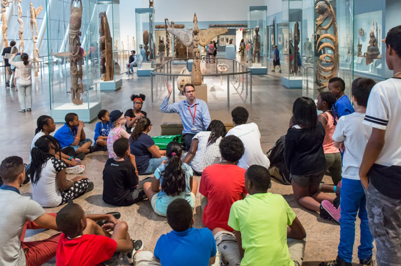 Students sit on the floor with their backs to the camera, watching a Met museum docent speak.