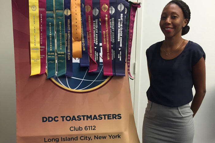 Shana Hitt, DDC Toastmasters club secretary, stands next to DDC toastmatsters sign with award ribbons placed on top.