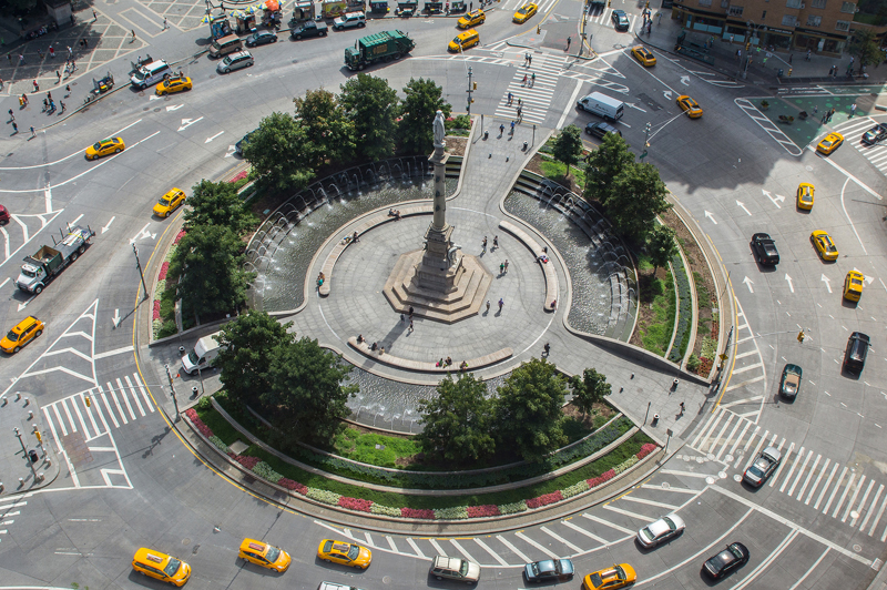 A birds eye view of Columbus Circle. A roundabout traffic intersection, with a statue, plantings, and benches in the center median.
