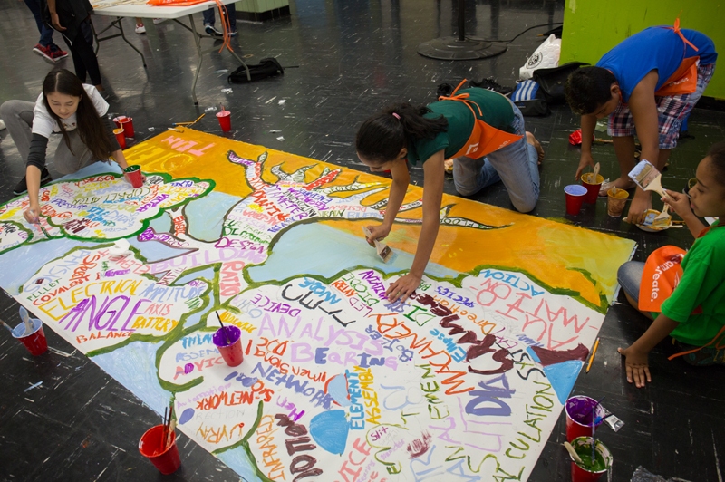 Students sit on the floor, painting on a large sheet of white paper.