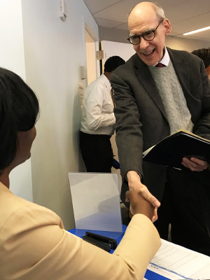  Older male wearing a suit jacket and holding a resume shakes the hand of a female professional sitting at a booth.