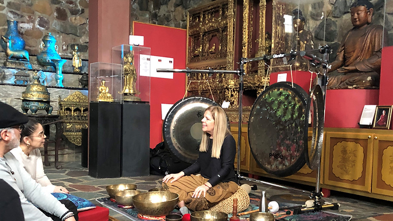 Blonde woman surrounded by meditation bowls with a man and a woman looking on as the receive instruction for meditation