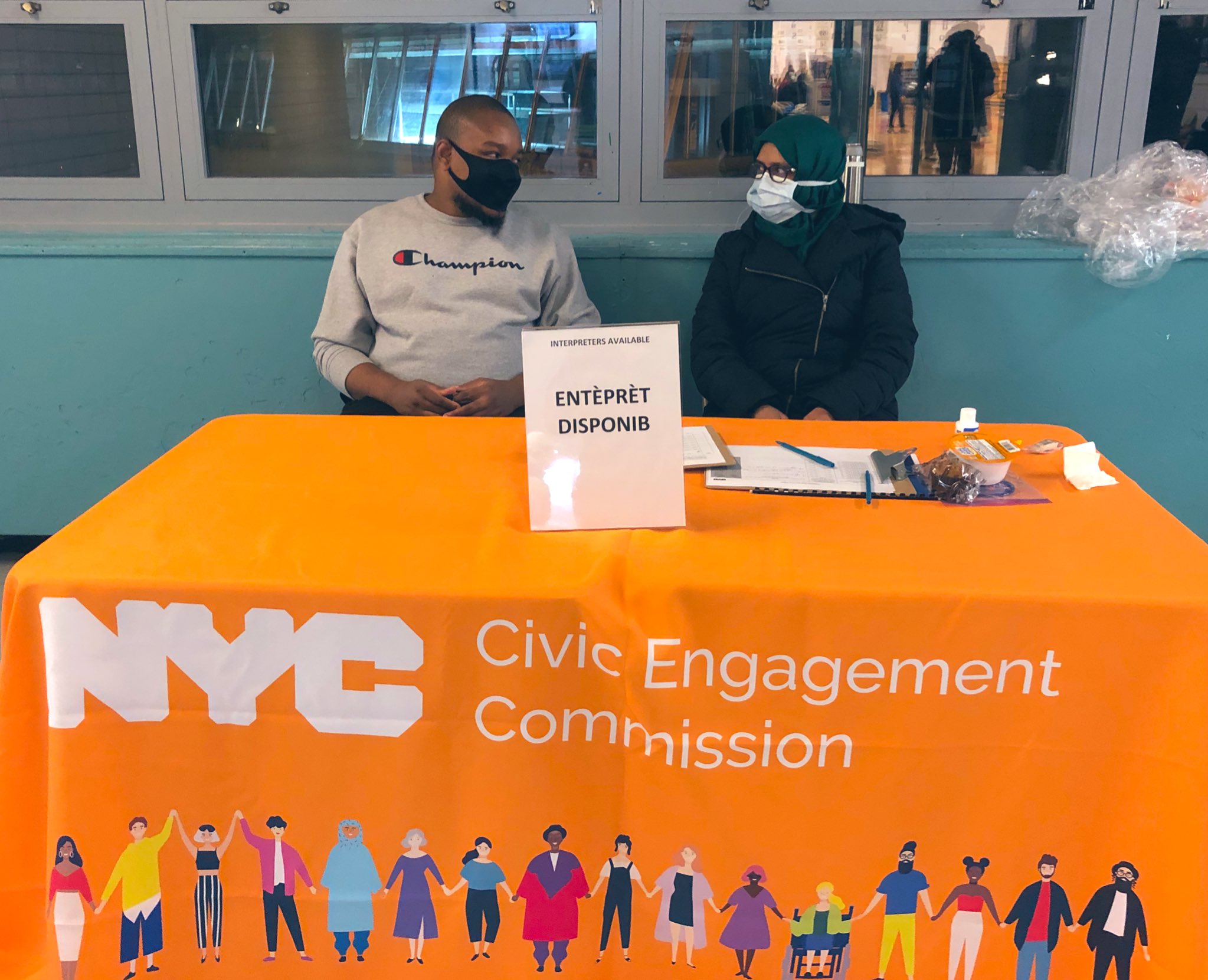 Two poll workers sitting at a table with orange table cloth with the NYC Civic Engagement Commission logo in front and a sign posted with Interpreters Available