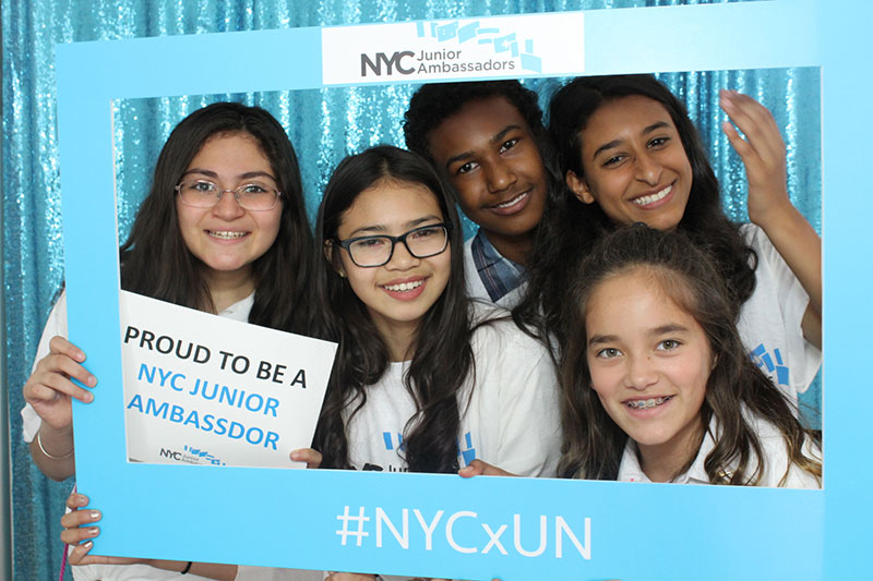 Four students standing close together, holding up a up a blue square frame that says: NYC Junior Ambassadors #NYCxNY
