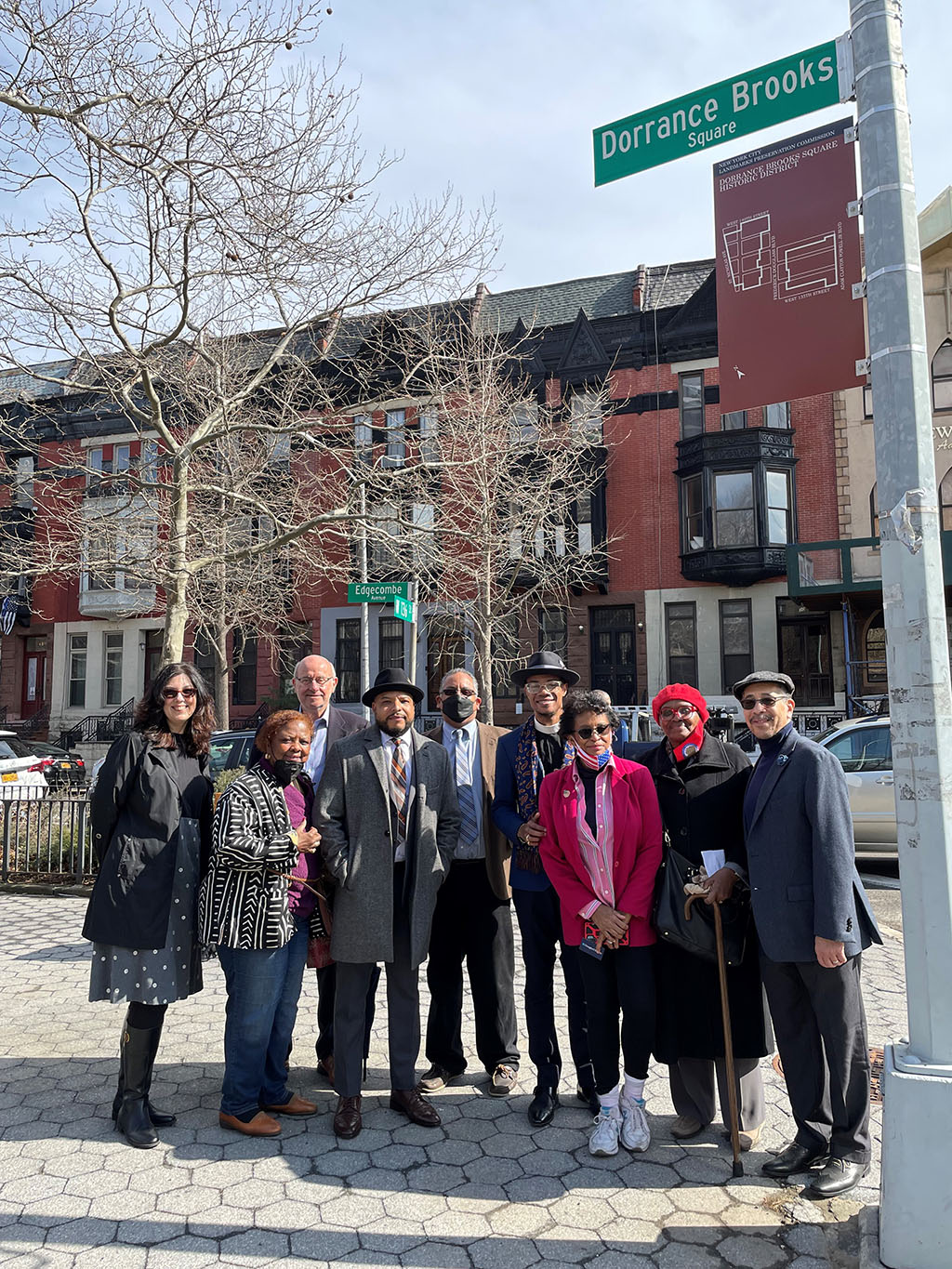 A group of people standing next to the Dorrance Brooks Sq Marker, part of the unveiling. The marker is a green nyc street sign with white text, Dorrance Brooks Square. Underneath the street sign is a marker sign that is burgundy, with with text and a outline of the square. 