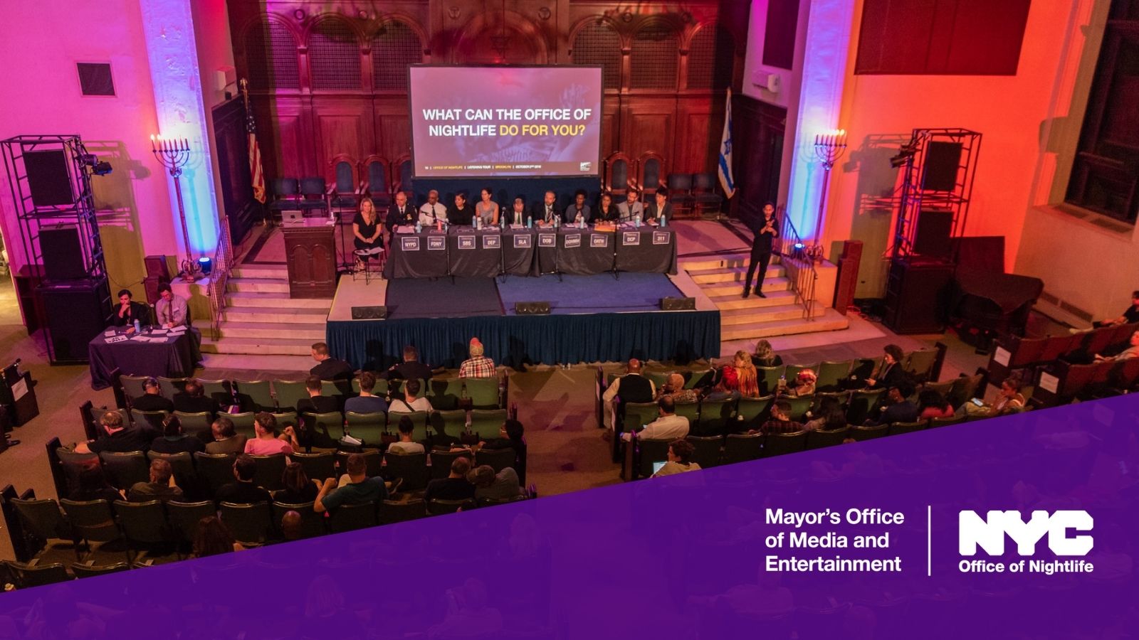 Overhead view of city officials on stage in front of a screen that reads “What can the Office of Nightlife Do For You” and community member sitting in the audience at the Office of Nightlife Town Hall