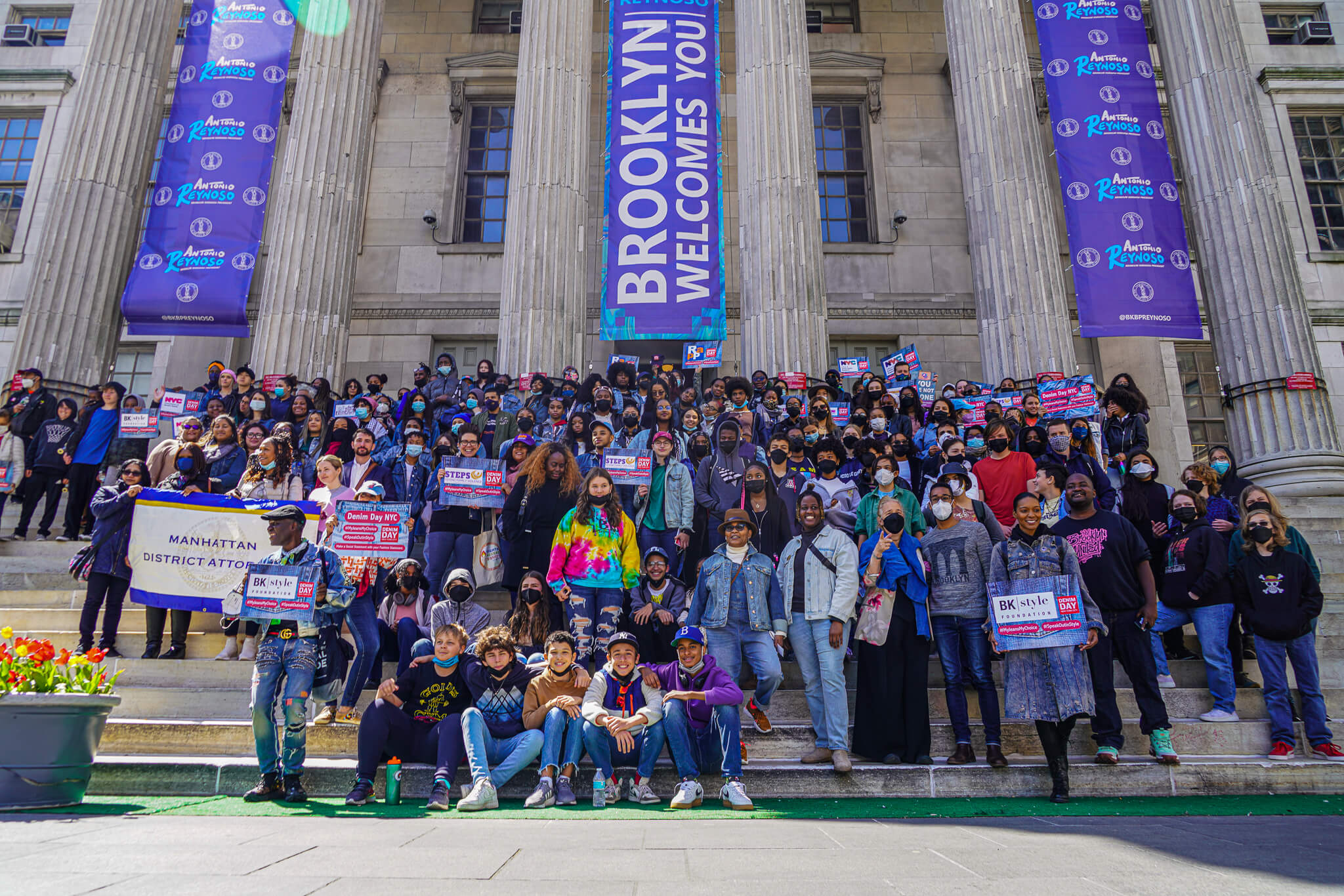 Posed image of approx. 100 people in denim outfits  holding Denim Day NYC signs standing on Brooklyn Borough Hall steps for Denim Day March