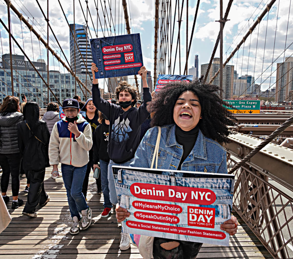 Marchers walking over Brooklyn Bridge in denim outfits holding Denim Day NYC signs including woman shouting.
