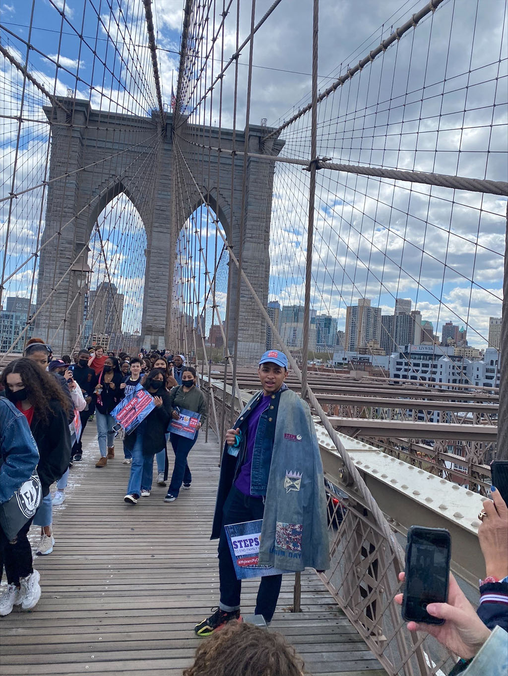 Image of posed male in long denim cape standing along side marchers holding Denim Day signs over Brooklyn Bridge to Manhattan.