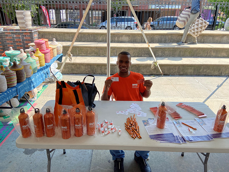  A male in orange t-shirt stationed behind table of flyers and giveaways in Brooklyn, NYC