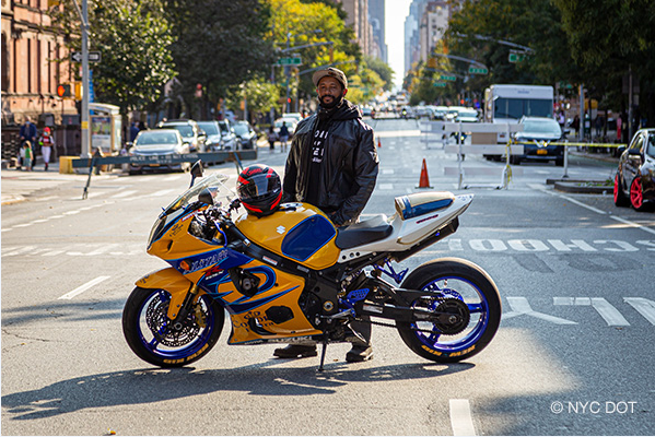 A man wearing motorcycle gear, stands beside a motorcycle parked on a street closed to traffic. His helmet rests on the motorcycle.