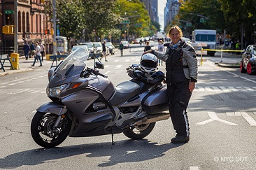 A woman wearing motorcycle gear, holding a helmet, stands beside a motorcycle parked on a street closed to traffic.