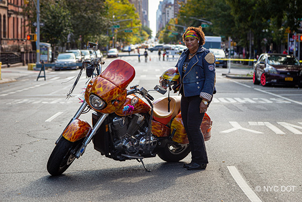 A woman holding a helmet stands beside a motorcycle, parked on a road closed to traffic.