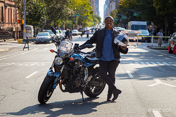 A woman wearing motorcycle gear, holding a helmet, stands beside a motorcycle parked on a street closed to traffic.