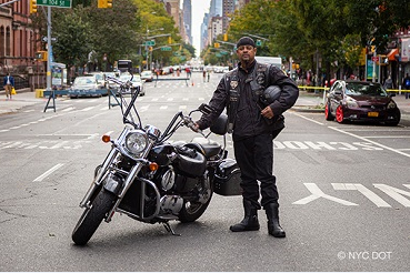A man wearing motorcycle gear, holding a helmet, stands beside a motorcycle parked on a street closed to traffic.