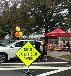 Picture of a Car Seat Fitting Area shows a white car parked near the curb and a red tent on the sidewalk. A yellow sign with black letters reads “Child Safety Seat Check”