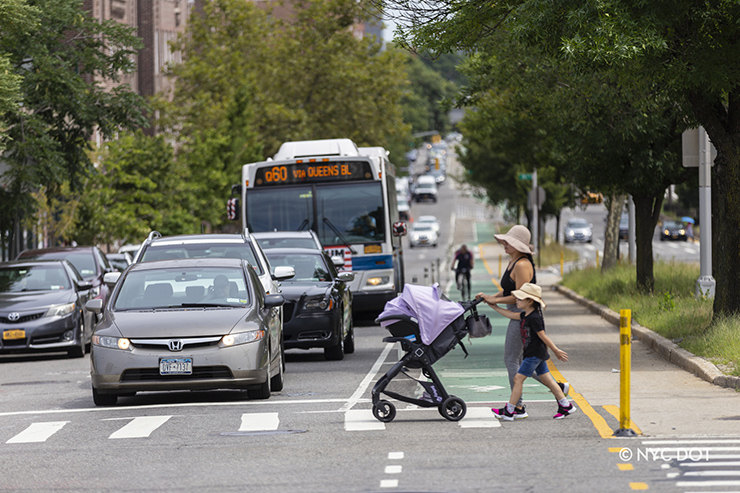 A woman and child push a stroller while walking in a crosswalk across a street on a sunny day in Queens. Cars and an MTA bus are stopped at the intersection, and a cyclist rides in a green bike lane towards the intersection.