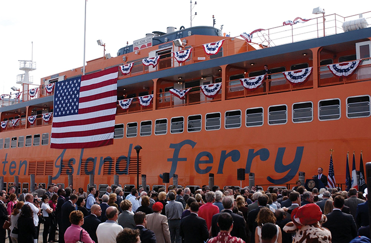 People gather on a dock looking at a large, orange Staten Island Ferry named for Guy V. Molinari. The ferry's decks are decorated with patriotic flags.