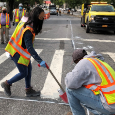 An NYC DOT worker wearing a yellow and orange safety vest sprays paint on a roadway, marking where the street will be redesigned.