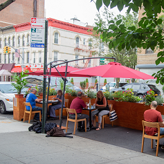 People sit at restaurant tables set up in a roadway dining area spaced six feet apart and separated from vehicles by a thick barrier.