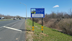 Blue Adopt a Highway sponsor sign on grassy area near highway. Sign Reads: 
 For a Cleaner Greener NYC 
 Site Available Call (718) 712-7563