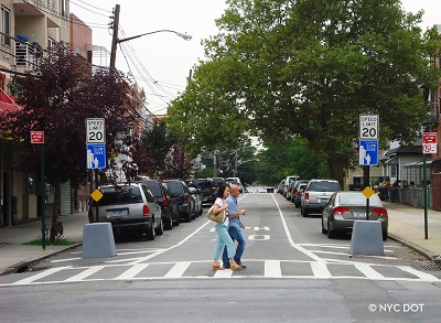 Two adults walk in the crosswalk, over a one-lane roadway with two white “Speed Limit 20” signs and two blue “Neighborhood Slow Zone” signs on either side of the road.