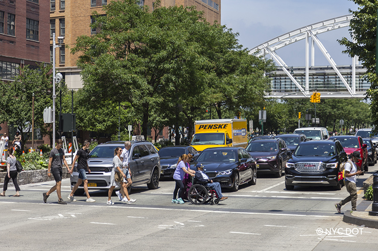 On a sunny day, pedestrians walk and push man sitting in a wheelchair through a crosswalk in Lower Manhattan while three lanes of cars and trucks wait at a traffic signal.