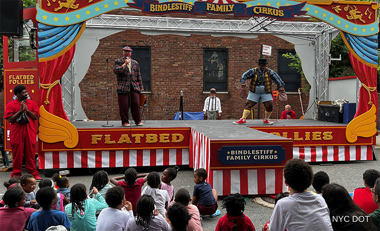 Two people juggle brightly colored clubs on a flatbed stage in a plaza.