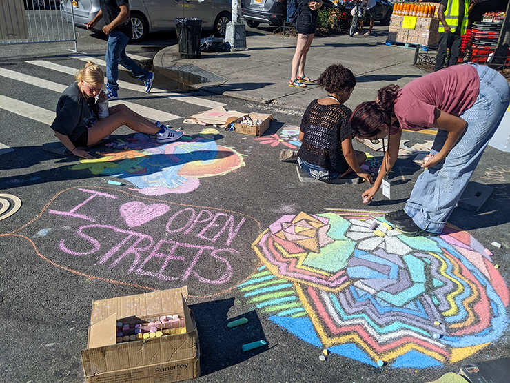 Three young women use chalk to make colorful designs on a roadway closed to vehicle traffic. One of the drawings on the roadway reads “I heart symbol Open Streets”.