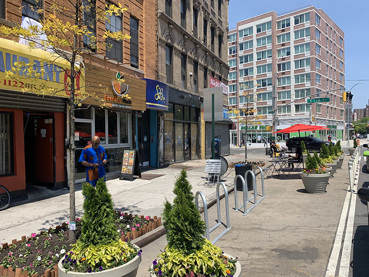 Curbside parking spaces have been converted into pedestrian space, filled with large planters, bike parking racks, tables, chairs and umbrellas. Between the vehicle travel lane and the pedestrian space is a line of flexible delineators and large planters.