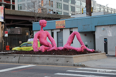 A large, pink sculpture of a person laying down is set up in a median between two lanes of traffic in Long Island City.