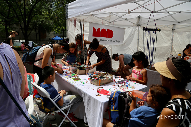Adults and children stand and sit around a folding table with arts and crafts setup. A white tent is setup behind the table, with a sign reading MAD